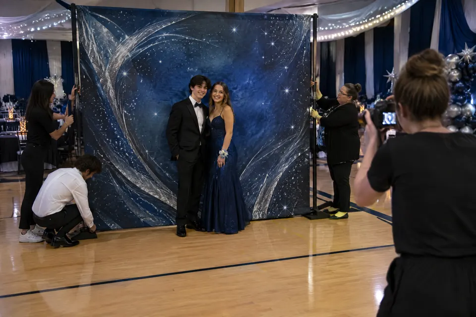 Students pose in front of a wide freestanding midnight-blue prom photo backdrop while volunteers secure the support frame.