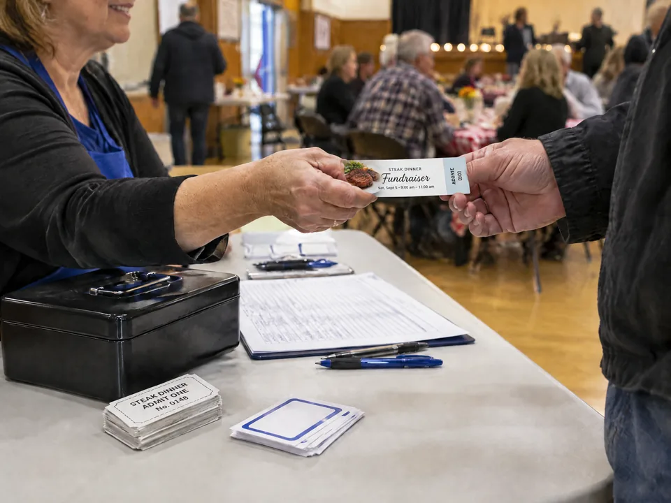 A volunteer hands a guest a long rectangular full-color steak dinner ticket with a narrow detachable stub.